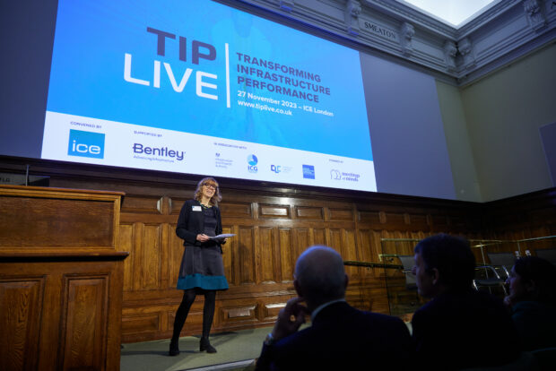 A woman stands on stage and addresses the audience at the TIP Live event. Behind her, a screen shows the TIP Live branding. Audience members are silhouetted in the foreground. 